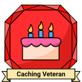 A stylized gemstone badge. It resembles a ruby and features a colored birthday cake. A ribbon reading Caching Veteran sits on the bottom.